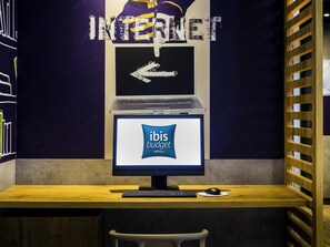 Un espace de travail avec un ordinateur portable, un clavier et une souris sur un bureau en bois, un écran d’ordinateur affichant « hôtels ibis budget », et un mur sur lequel figure un grand panneau « INTERNET » ainsi qu’une flèche pointant vers la gauche.