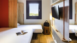 A hotel room with two beds, a window with a view, a TV mounted on the wall, and a small stool. - Ibis budget Stein Maastricht (Stein)