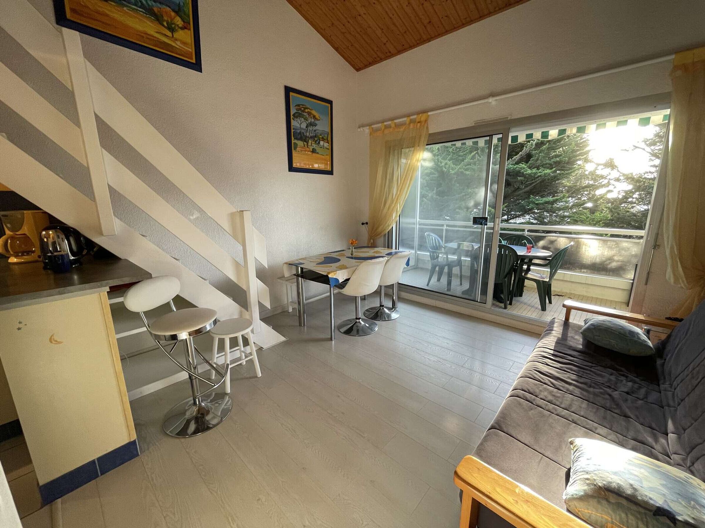 Ein modernes Wohnzimmer mit einer Couch, einem Esstisch mit Stühlen, einer Küchenzeile inklusive Kühlschrank und einer großen Glasschiebetür, die auf eine Terrasse im Freien führt.