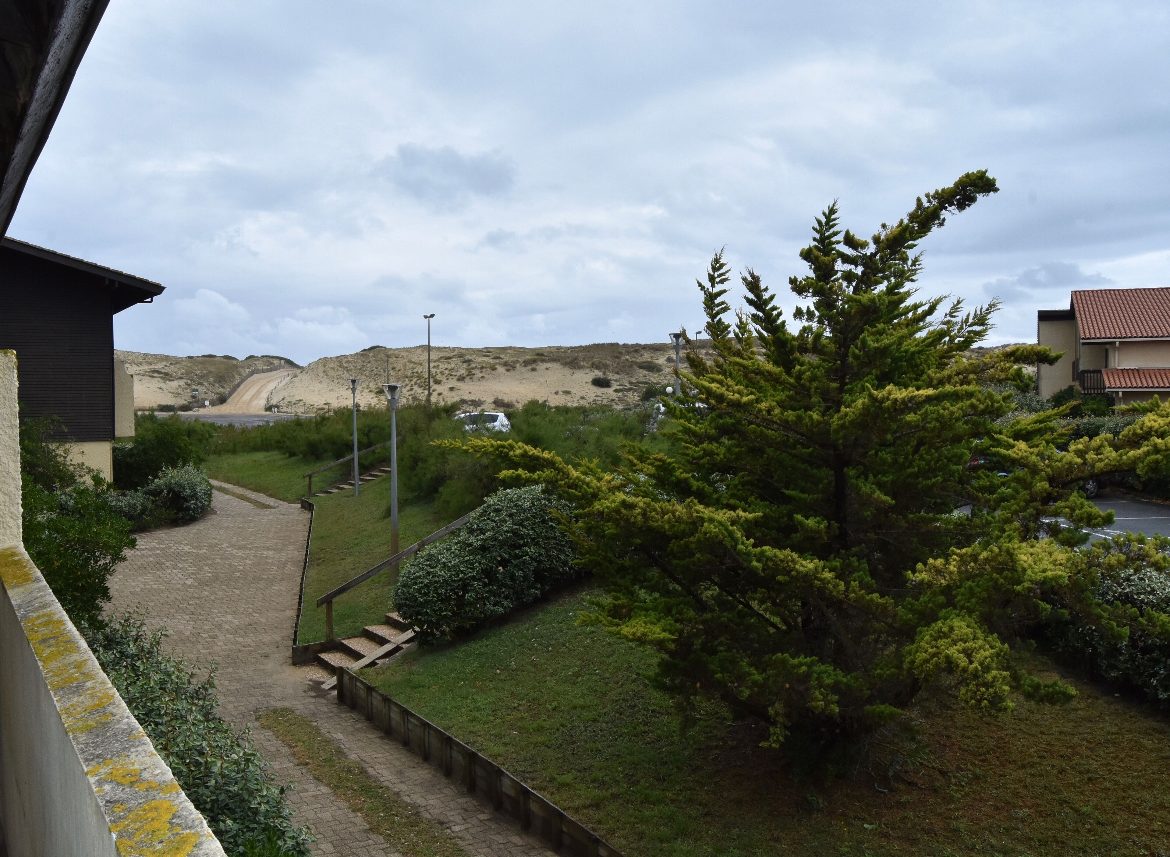 Un chemin menant à un bâtiment d’où l’on peut admirer une colline, ainsi qu’à un parking où sont garées des voitures.