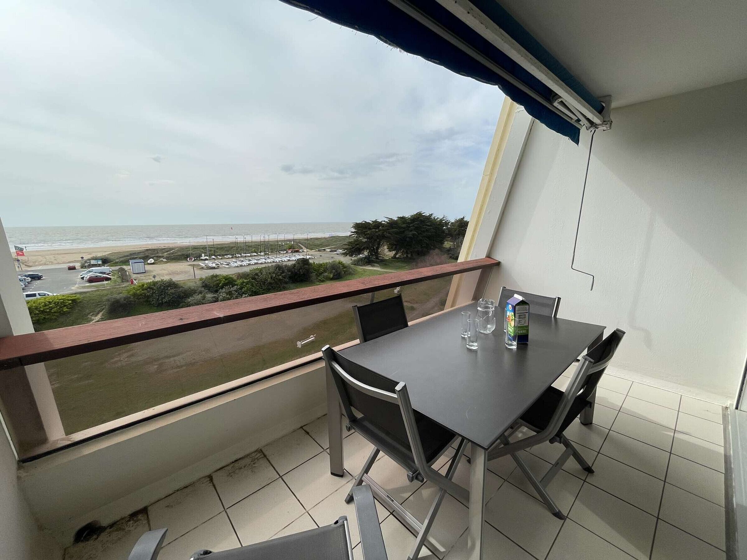 Een balkon met een tafel en stoelen, met uitzicht op een strand en een parkeerplaats.