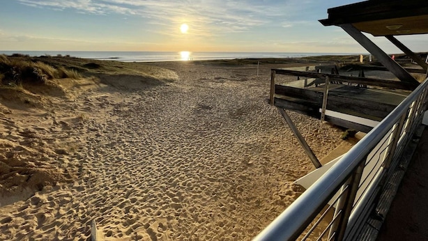 Ein Strand mit einer Holzpromenade, die zum Wasser führt, bewölkter Himmel und eine untergehende Sonne in der Ferne.