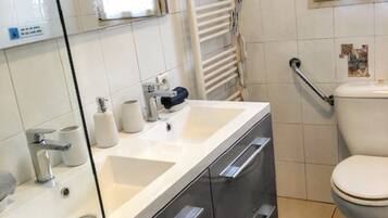 Un baño con lavamanos blanco, espejo y retrete. Hay un perchero y una alcachofa de ducha en la pared.