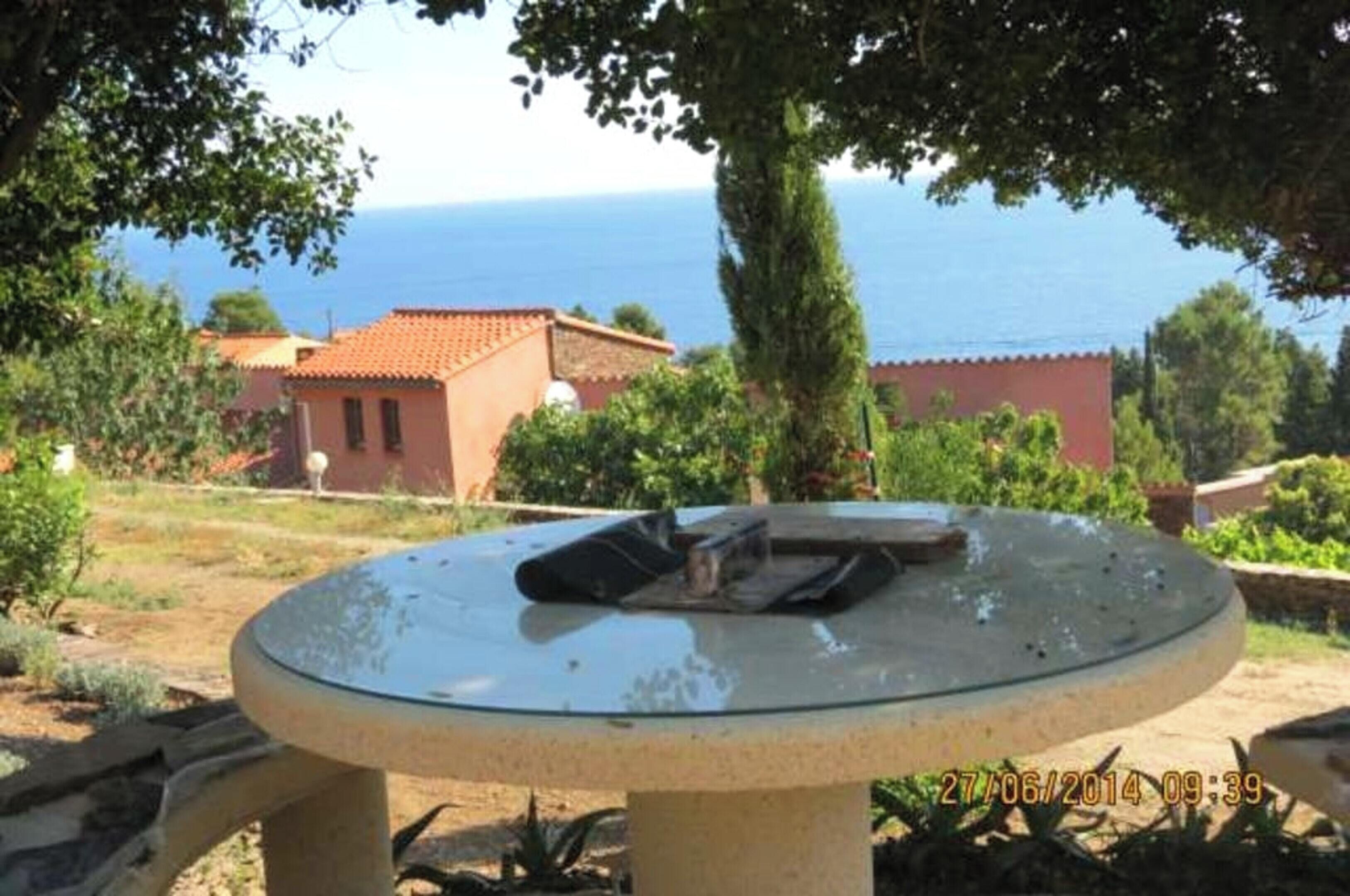 Un espacio de estar al aire libre con forma circular, con una mesa y sillas, rodeado de follaje y edificios con tejados rojos, con vista a un cuerpo de agua.