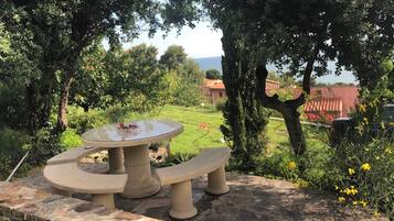 Una mesa de piedra para picnic con bancos, rodeada de vegetación y con vista a una casa al fondo.
