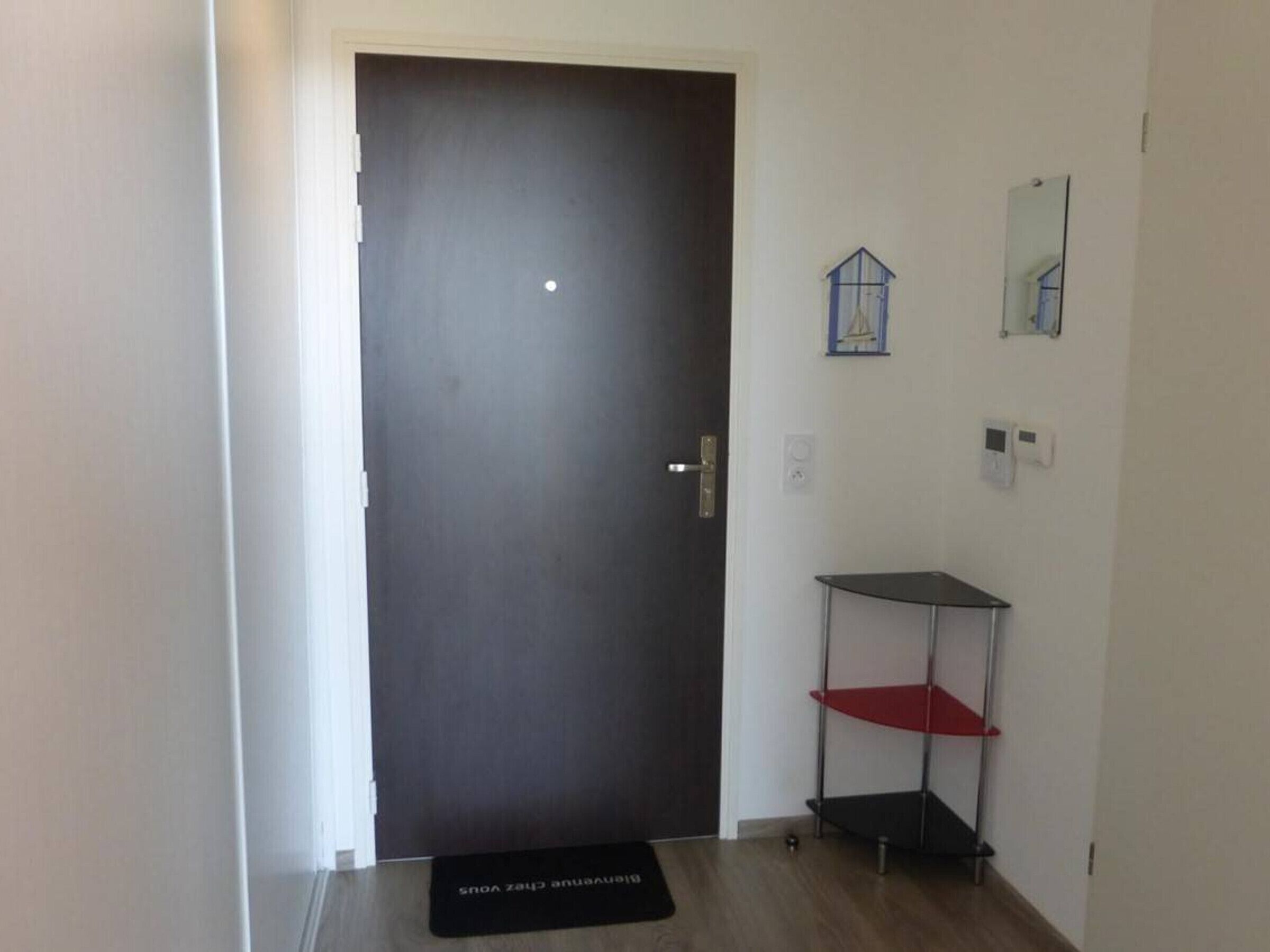 Una puerta oscura cerrada con un picaporte plateado, un mueble estante pequeño con repisas rojas y negras, y un felpudo negro con texto blanco en el suelo.