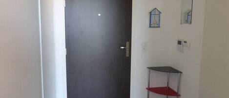 Una puerta oscura cerrada con un picaporte plateado, un mueble estante pequeño con repisas rojas y negras, y un felpudo negro con texto blanco en el suelo.