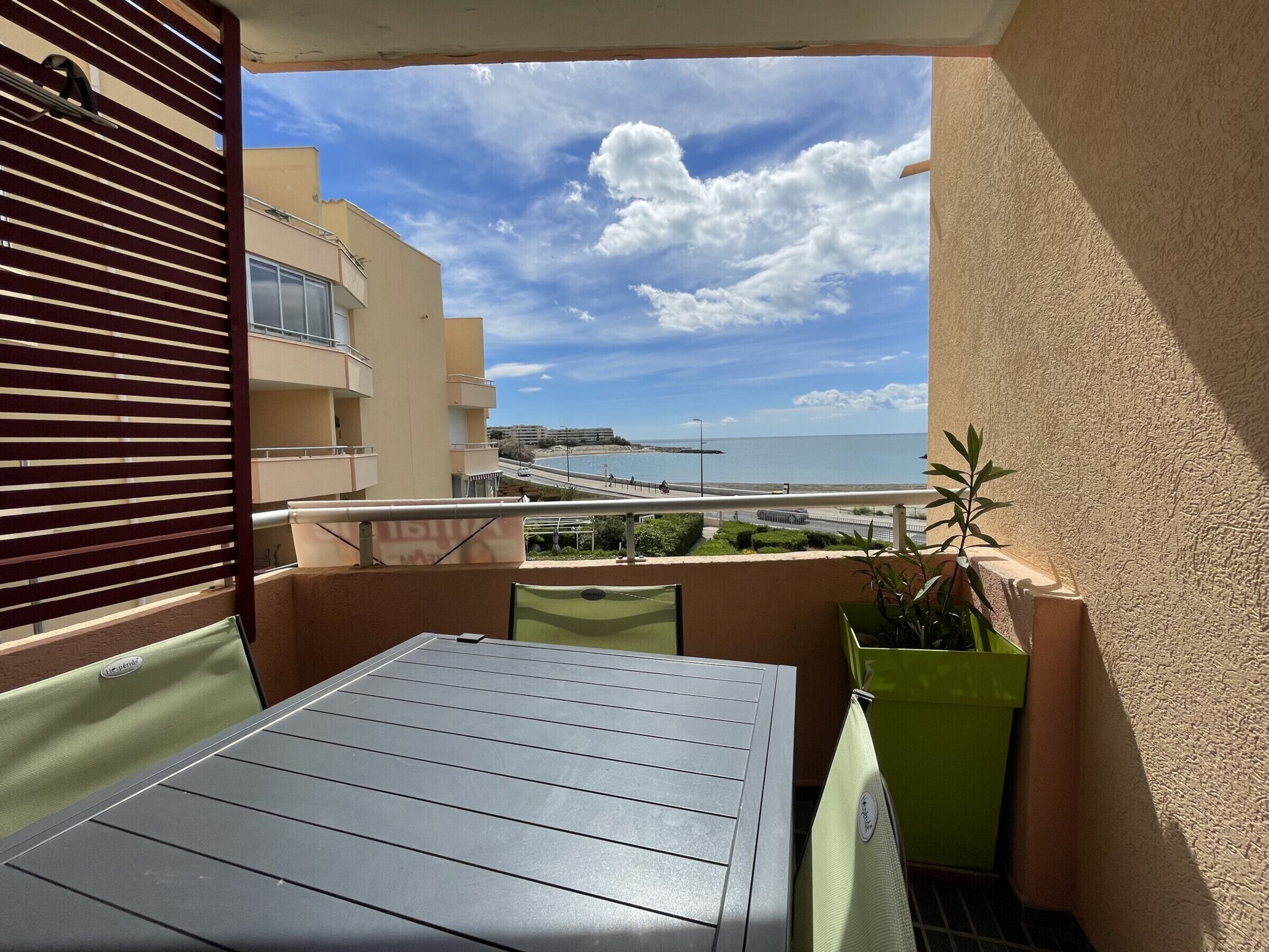 A balcony with a table and chairs, a potted plant, and a view of a beach and buildings.