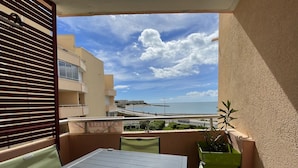 Un balcón con mesa y sillas, una planta en maceta y vista a una playa y edificios.