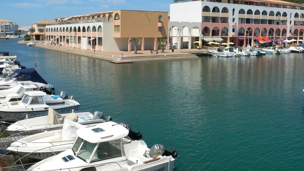 Un puerto deportivo con barcos amarrados, un edificio con ventanales arqueados y un cielo despejado.