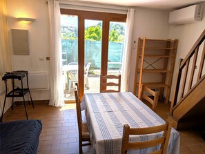 Huoneessa on pöytä, tuoleja, pieni jakkara, patteri, ikkuna verhoilla, portaikko ja näkymä ulos.