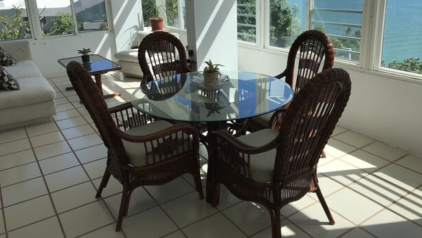 A dining area with a glass table surrounded by wicker chairs, a sofa, and a view of the sea. - El Encanto de Naturaleza y Romantic! Apartment 2 (Fajardo)