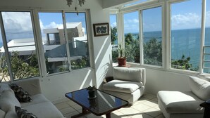 A sunlit room with a view of the sea, featuring a sofa, a table with a plant, and a potted plant on a windowsill. - El Encanto de Naturaleza y Romantic! Apartment 2 (Fajardo)