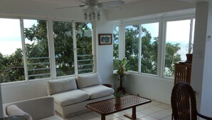 A room with a sofa, a small table, and a chair. There are large windows with a view of trees and the sea. - El Encanto de Naturaleza y Romantic! Apartment 2 (Fajardo)