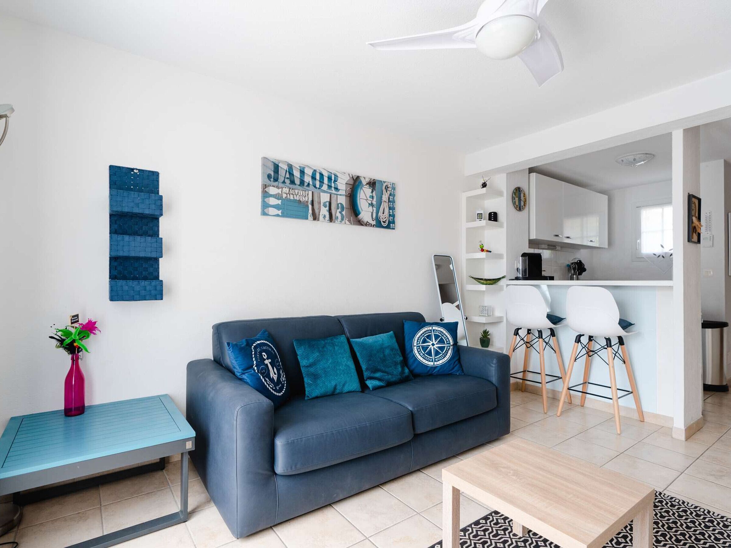 A modern living room with a dark blue sofa, a small table, and a coffee table. There is a kitchen area with white cabinets and a dining space with white chairs.