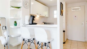 Une cuisine de taille réduite avec des meubles blancs, une machine à café et une horloge au mur. On y trouve deux chaises blanches aux pieds noirs.