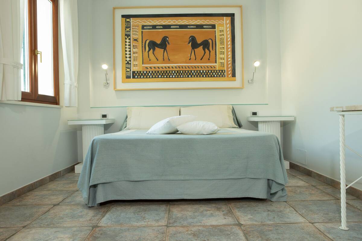 A bedroom with a bed, two bedside tables, a framed artwork of horses, and a window with curtains.