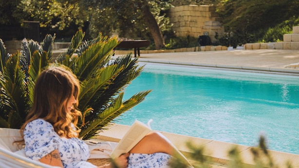 Une personne se détend au bord d’une piscine, un livre à la main, entourée de verdure et d’un mur de pierres.