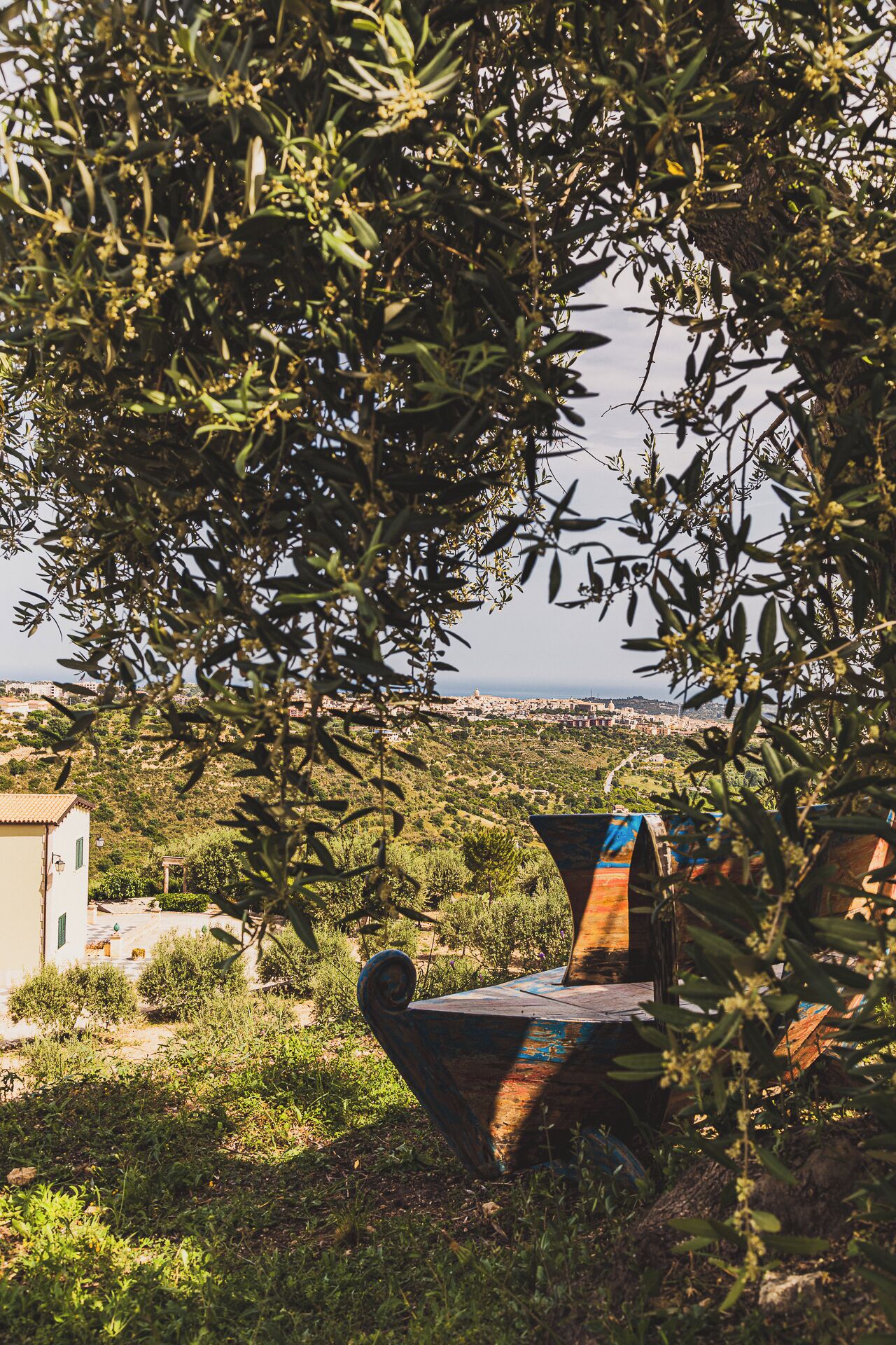 A rustic outdoor seating area with a metal bench, surrounded by olive trees and greenery, overlooking a coastal town.