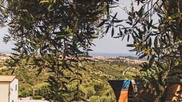 A rustic outdoor seating area with a metal bench, surrounded by olive trees and greenery, overlooking a coastal town.