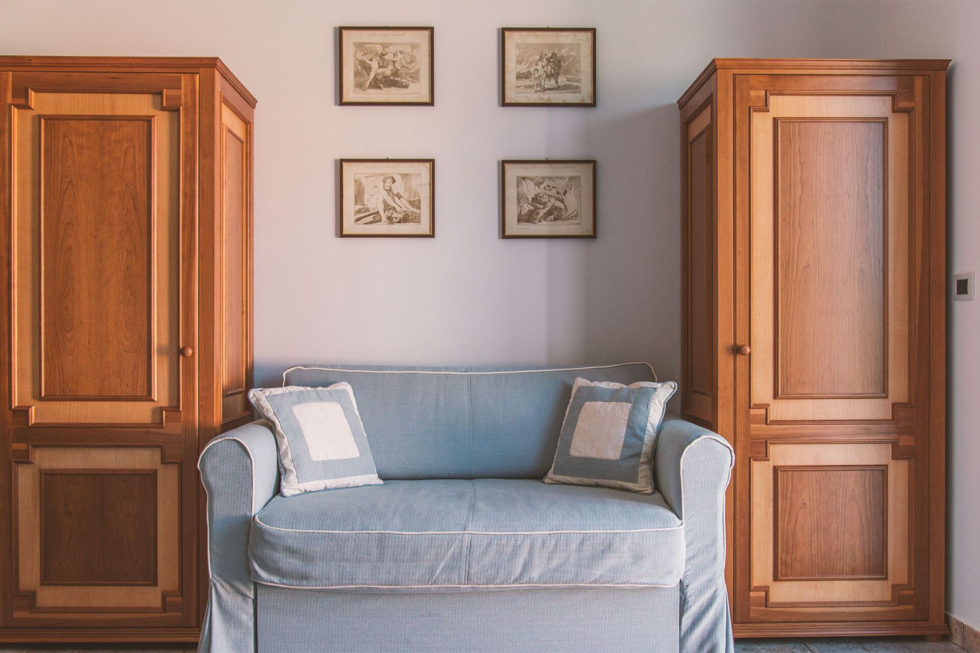 A room with a light blue sofa, two matching pillows, and three framed pictures on the wall. There are two wooden cabinets on either side of the sofa.