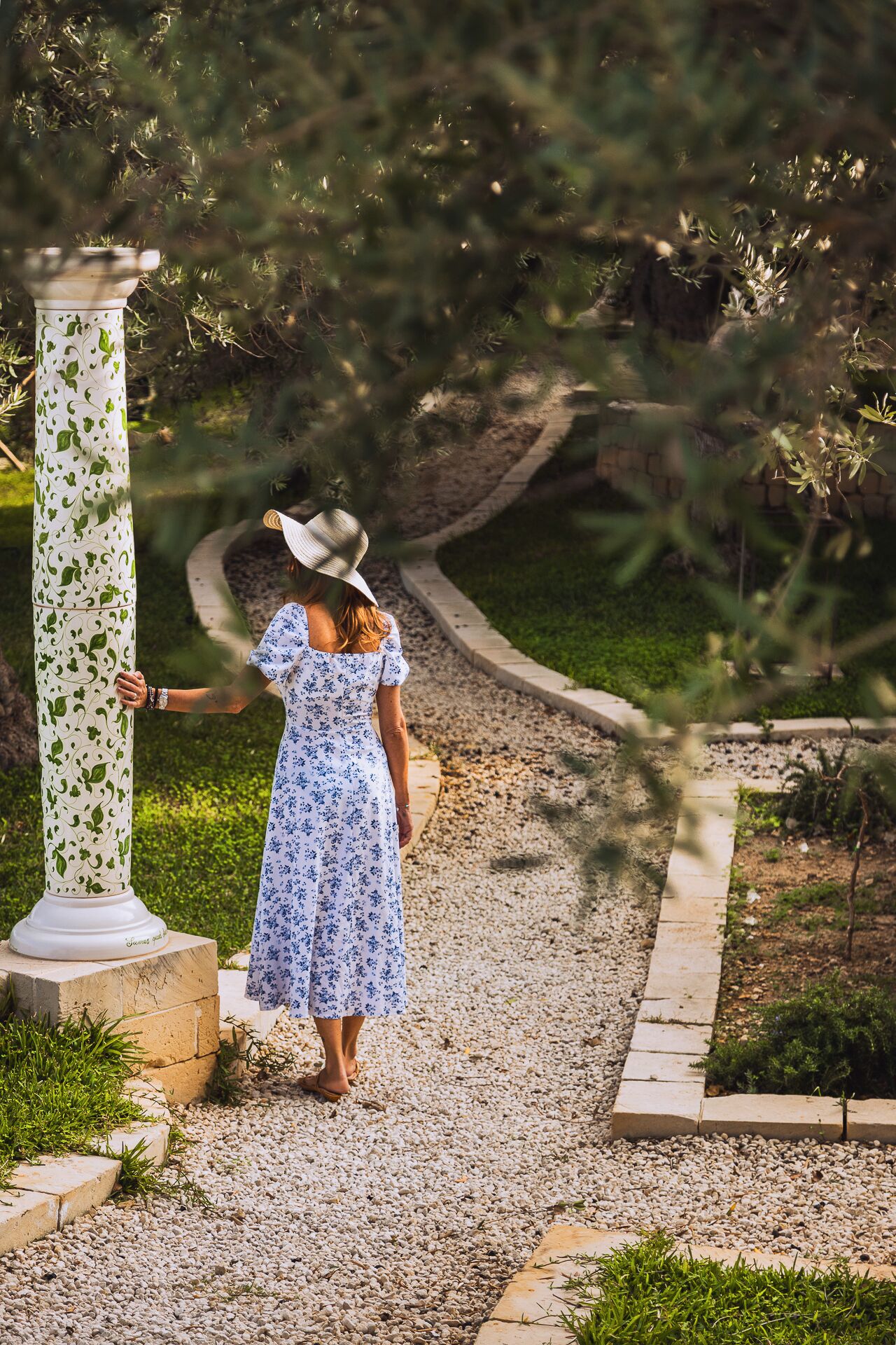 Una mujer con vestido y sombrero floral, de pie junto a una columna con decoración en la parte superior.