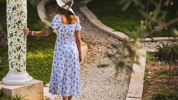 Una mujer con vestido y sombrero floral, de pie junto a una columna con decoración en la parte superior.