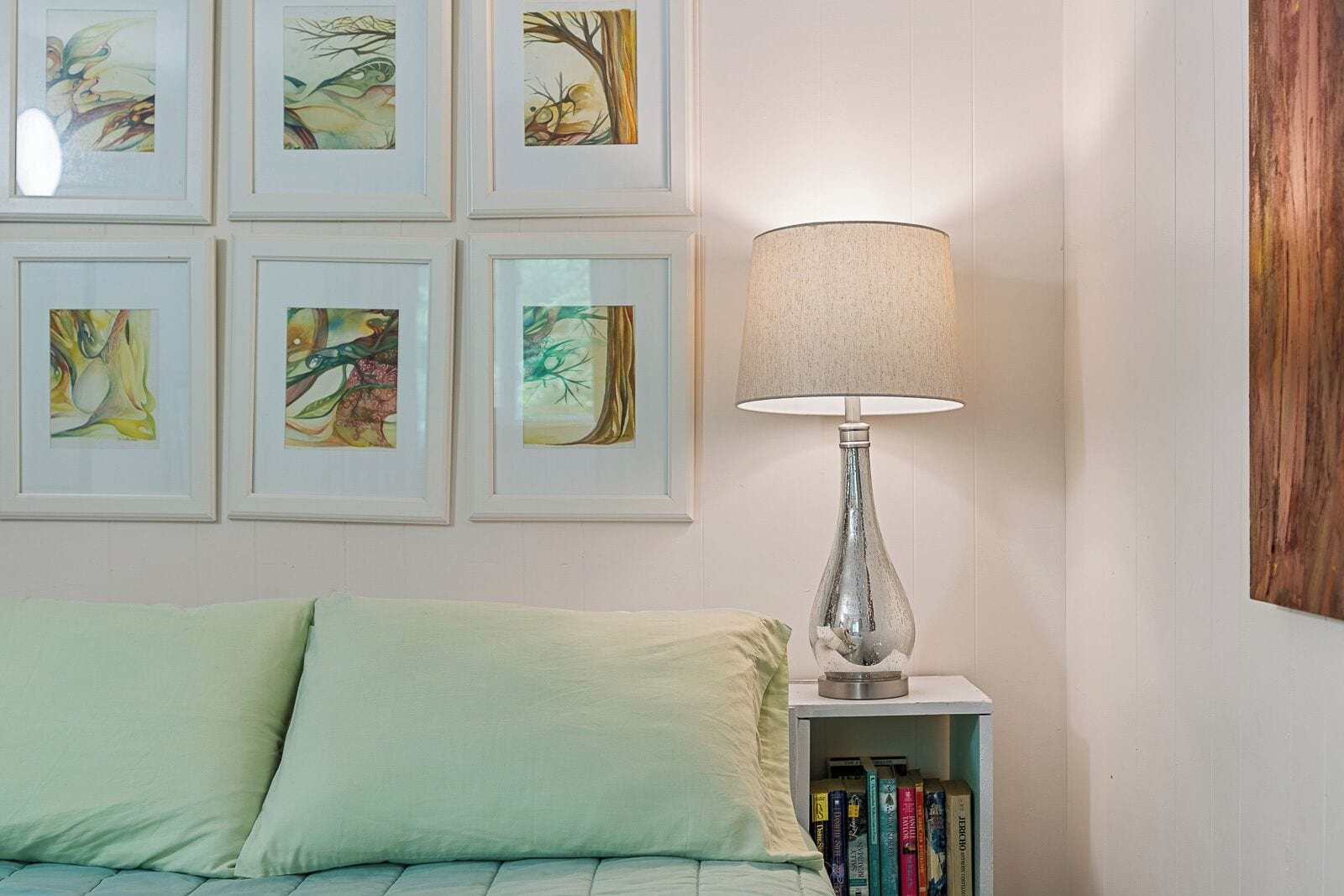 Una habitación con una cama, una mesita de noche con lámpara, y seis cuadros enmarcados en la pared.