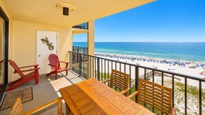 A balcony with a view of the beach, a wooden table, two red chairs, and a door with a lizard decoration.