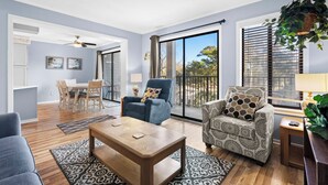 Amplia sala de estar con un gran ventanal corredizo a un balcón, un comedor con mesa y sillas, y una zona de estar acogedora con un sillón estampado y un sofá azul.