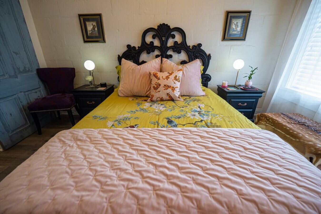 Una cama con cubrecama floral, cabecera, dos mesitas de noche con lámparas, una silla y una ventana con persianas.