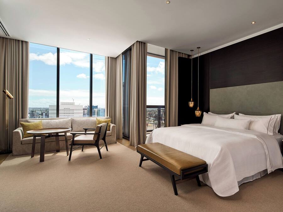 A modern hotel room with a large bed, a seating area with a table and chairs, and a view of the city through large windows.