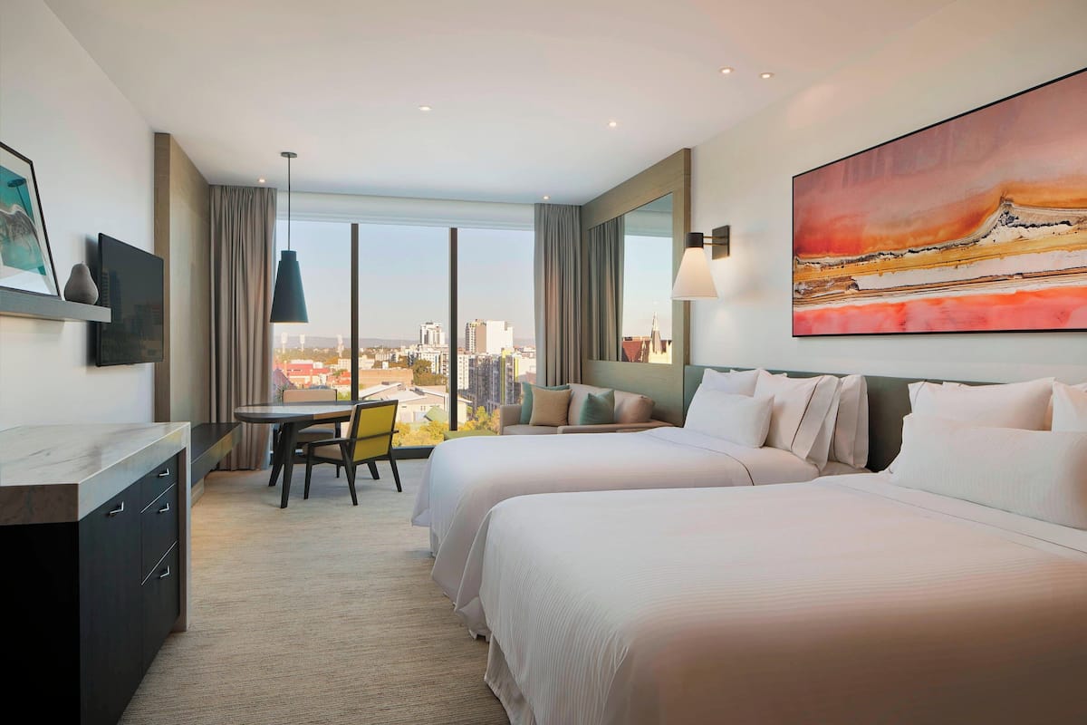 Quarto de hotel com duas camas, uma escrivaninha, uma cadeira e uma janela grande com vista para a cidade.
