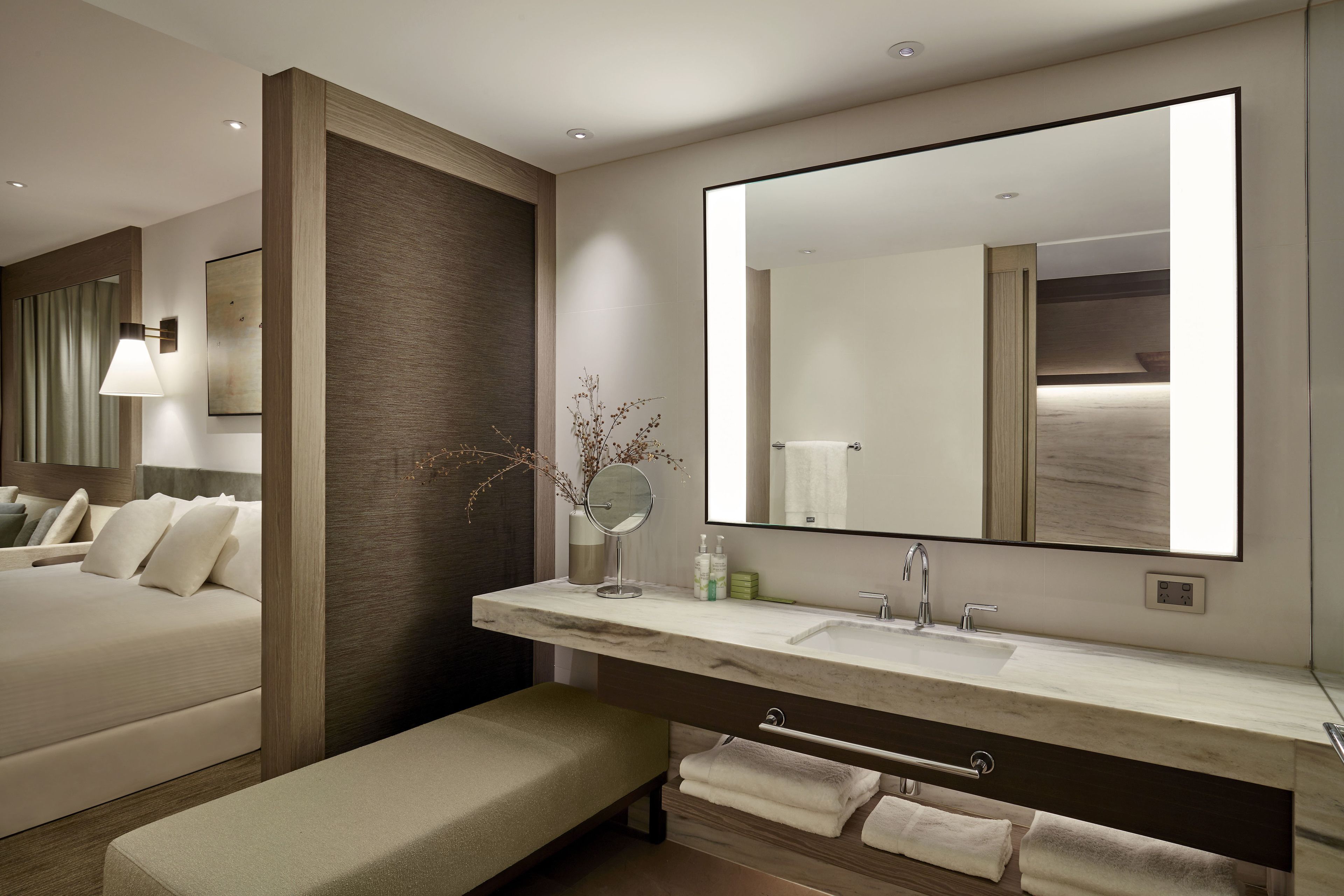 A modern hotel bathroom with a large mirror, dual sinks, and a bench.