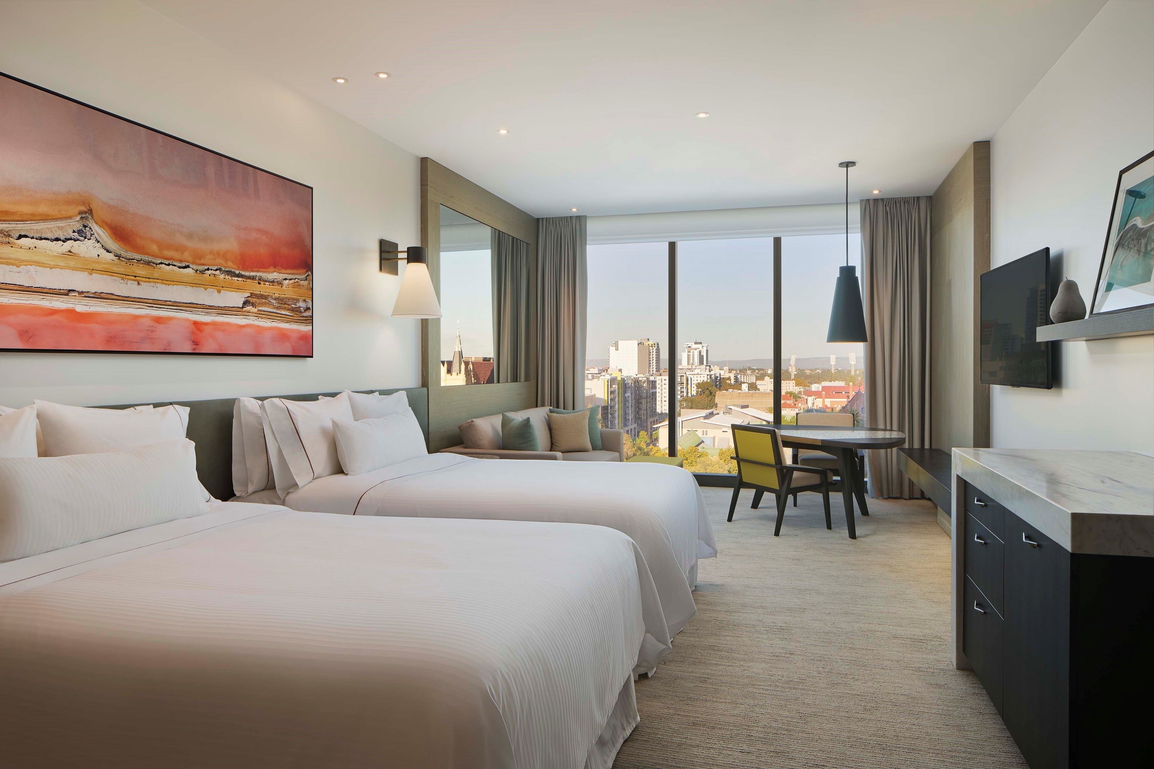 A hotel room with two beds, a dining table, a TV, and a large window with a city view.