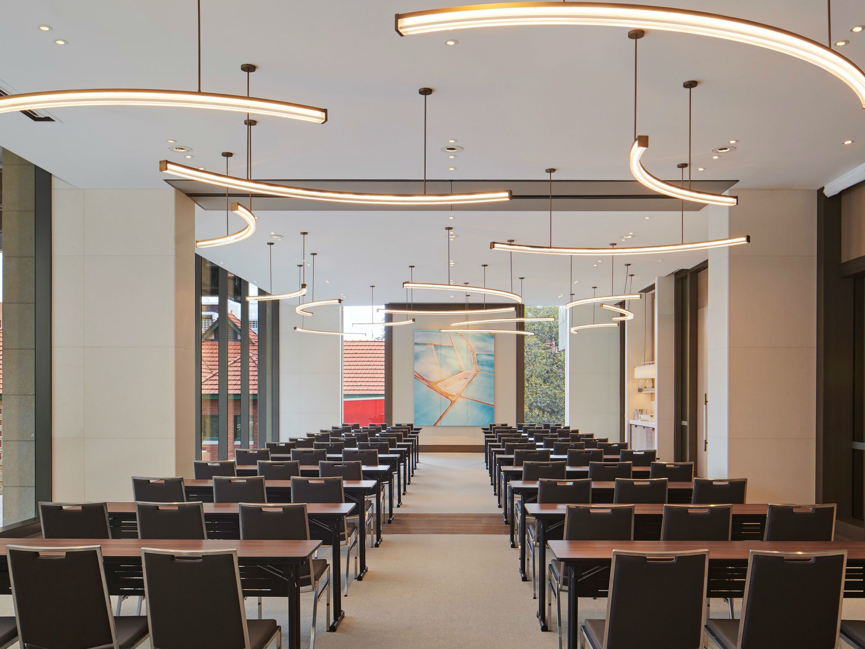 A conference room with rows of tables and chairs, modern pendant lighting, and a large abstract painting on the wall.