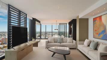 A modern living room with a large flat-screen TV, a sofa, a coffee table, and a city view through floor-to-ceiling windows.