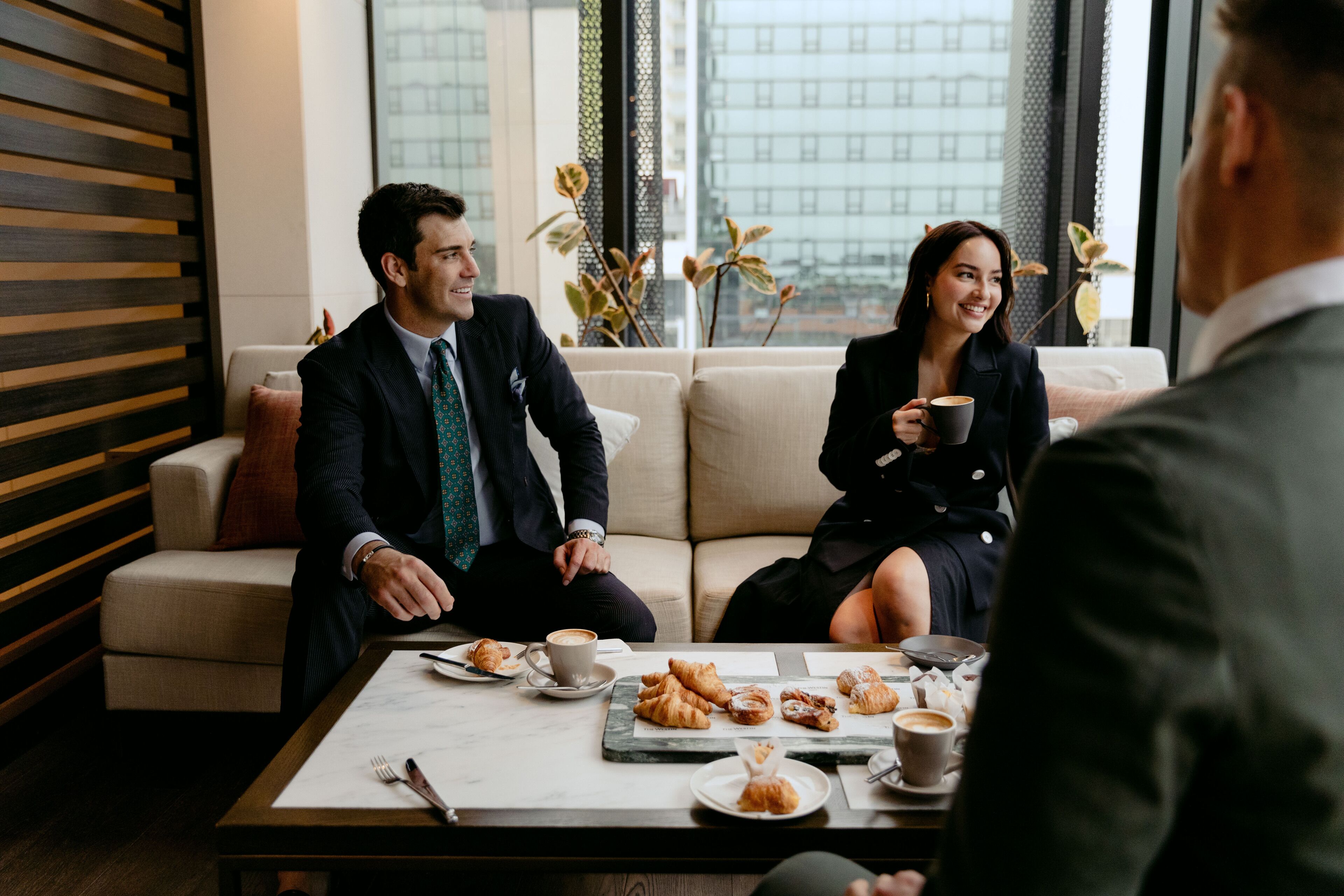 Une salle de réunion moderne, dotée d’un canapé, d’une table basse avec des pâtisseries et des tasses, et offrant une vue sur la ville grâce à de grandes fenêtres.