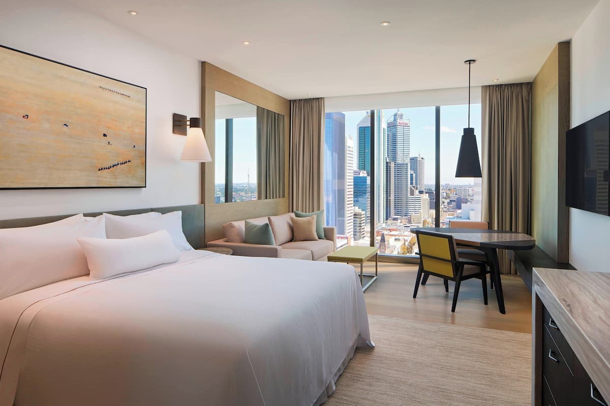 Quarto de hotel moderno com uma cama grande, um sofá, uma mesa de jantar e vista para a cidade.