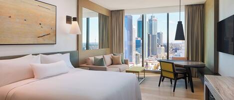 A modern hotel room with a large bed, a sofa, a dining table, and a city view.