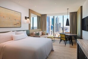 A modern hotel room with a large bed, a sofa, a dining table, and a city view.
