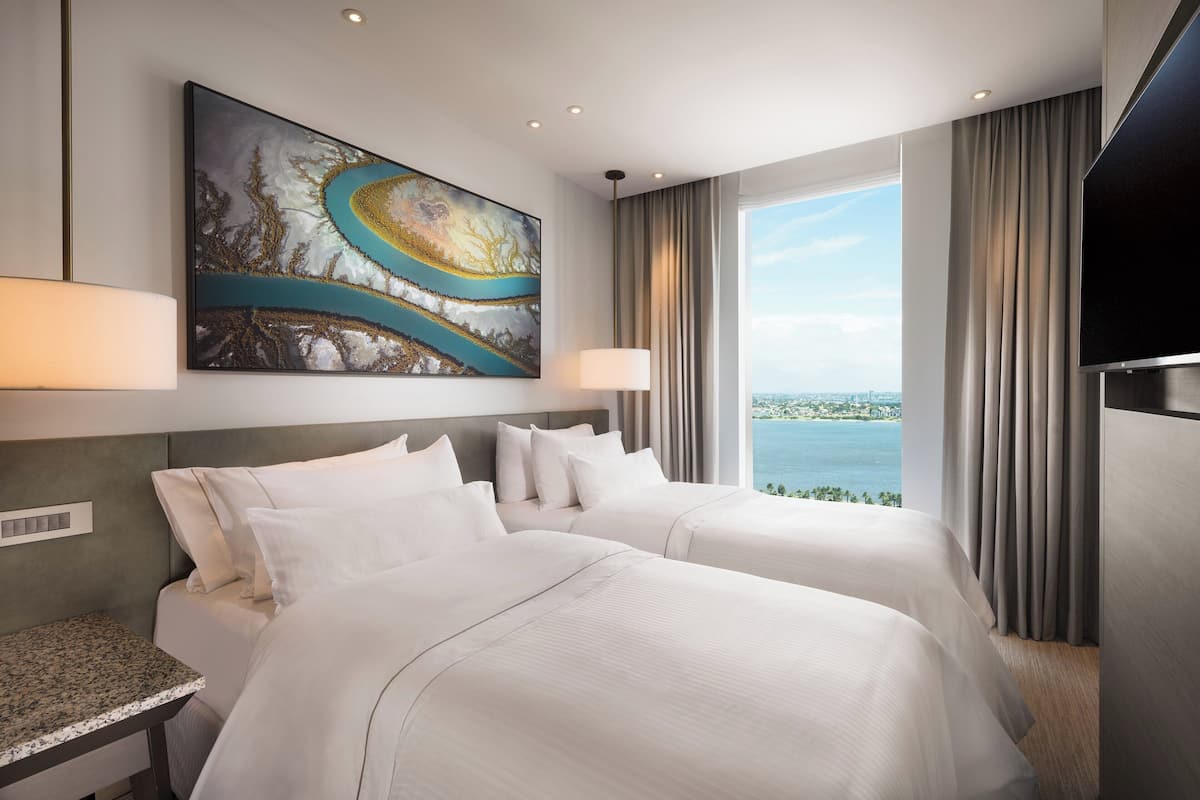 Quarto de hotel com duas camas, uma janela grande com vista, um quadro na parede e uma televisão de tela plana.