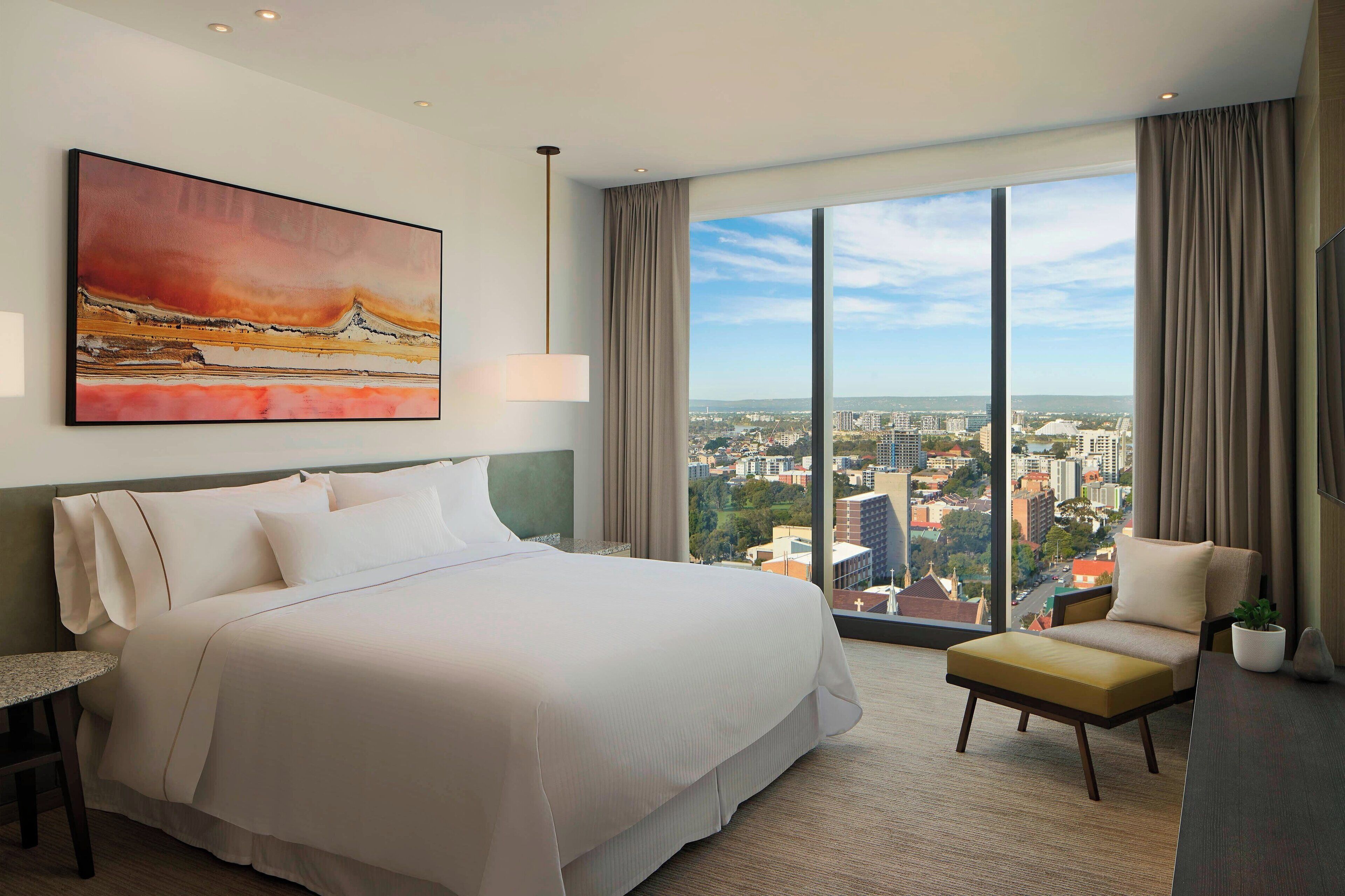 Habitación de hotel con una cama grande, una silla, un escritorio y vistas a la ciudad a través de amplias ventanas.