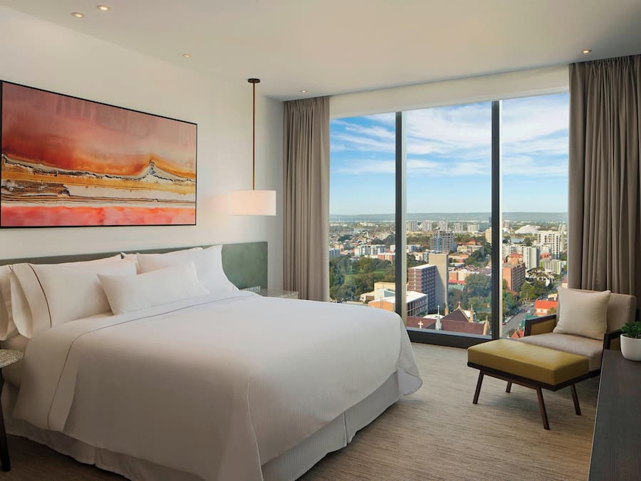 Habitación de hotel con una cama grande, una silla, un escritorio y vistas a la ciudad a través de amplias ventanas.