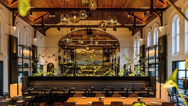 A restaurant with a long wooden table, black chairs, and a high ceiling with hanging lights. - The Westin Perth (Perth)