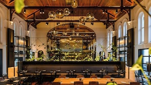 A restaurant with a long wooden table, black chairs, and a high ceiling with hanging lights.
