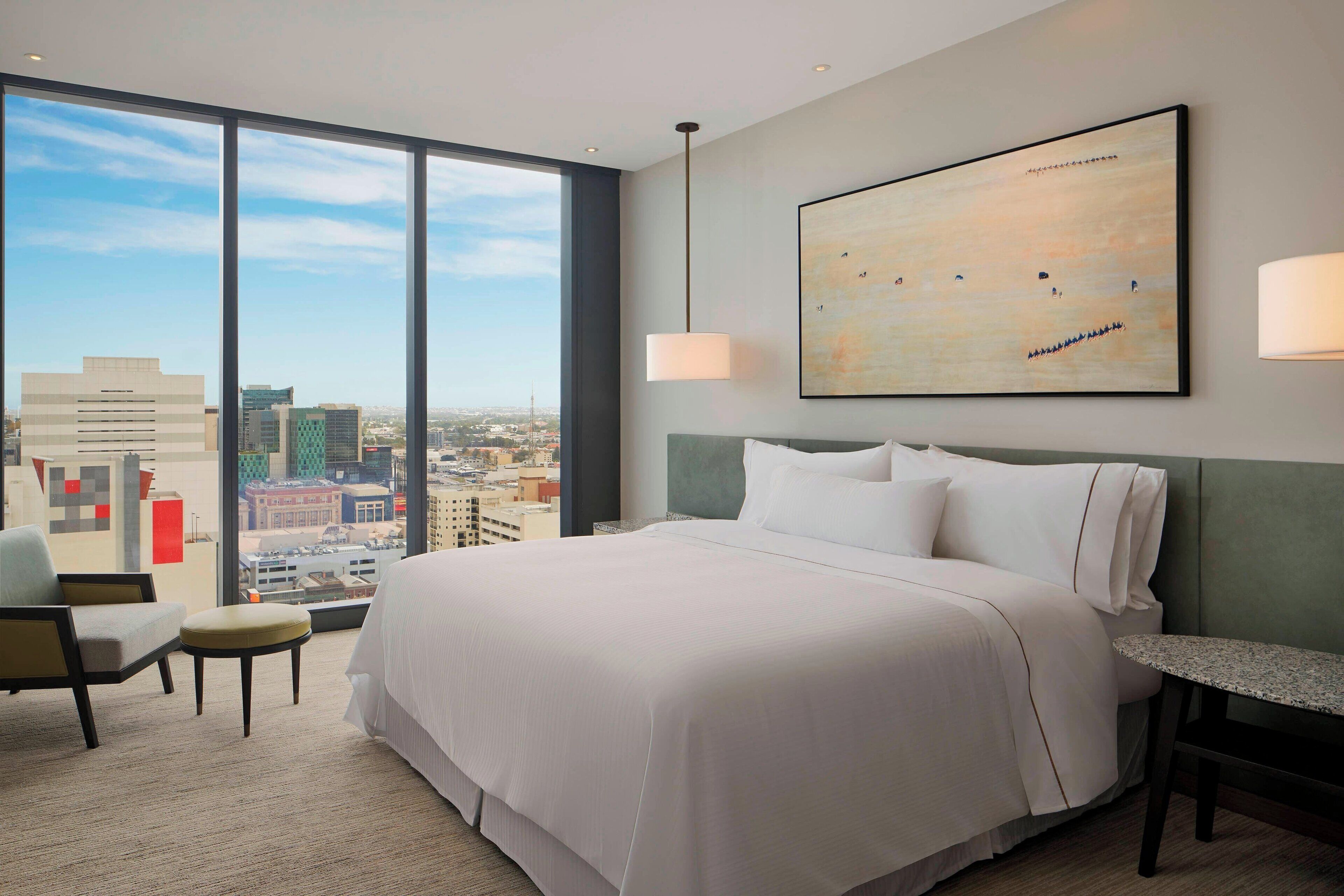 A hotel room with a large bed, a chair, a small table, and a city view through large windows.