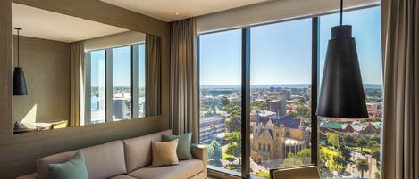 A modern hotel room with a large window offering a city view, a sofa, a dining table, and a hanging pendant light.