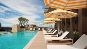 A rooftop pool area with lounge chairs and striped umbrellas, surrounded by wooden deck and a view of the city skyline.