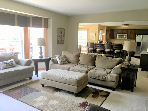 A living room with a sofa set, a coffee table, and a rug. There is a kitchen area with cabinets and a refrigerator in the background.